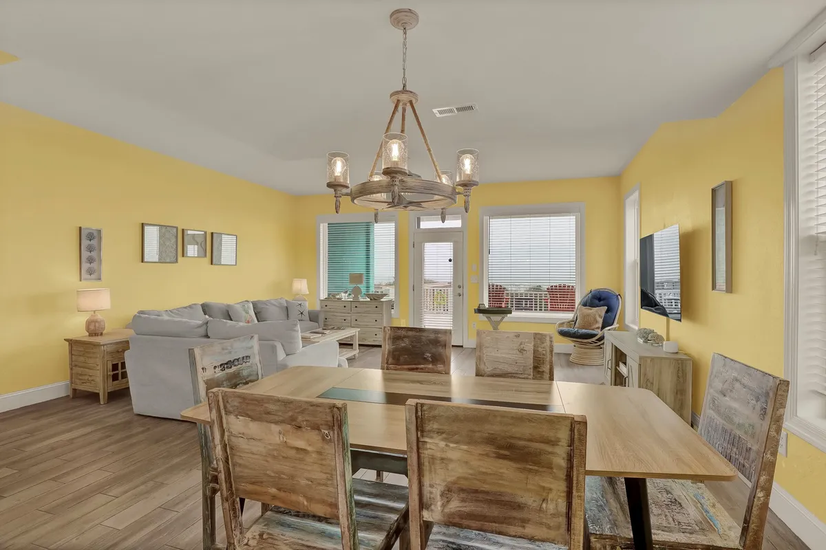 A cheerful dining space with a central dining table, a comfortable sofa nearby, an elegant chandelier, and vibrant yellow walls. The scene exudes warmth and hospitality, ideal for family dinners or casual lunches after a day at the beach.
