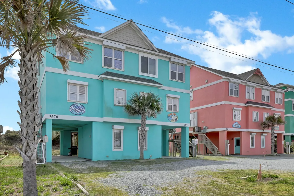 The image features two brightly colored buildings—one turquoise, one pink—framed by palm trees and street signs, capturing a lively, welcoming neighborhood vibe steps from the beach. The scene hints at the convenience of amenities (like EV charging) within a vibrant, walkable beach community.