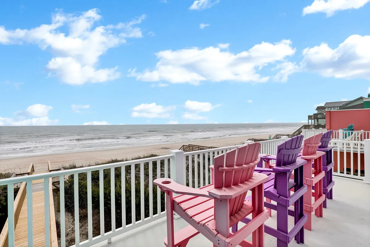 A panoramic view from a deck or patio with colorful Adirondack chairs overlooking the ocean and sandy beach, emphasizing direct beach access and communal outdoor seating. The tranquil scene reflects opportunities for sunbathing, relaxed conversations, and group meals with a stunning seaside backdrop.