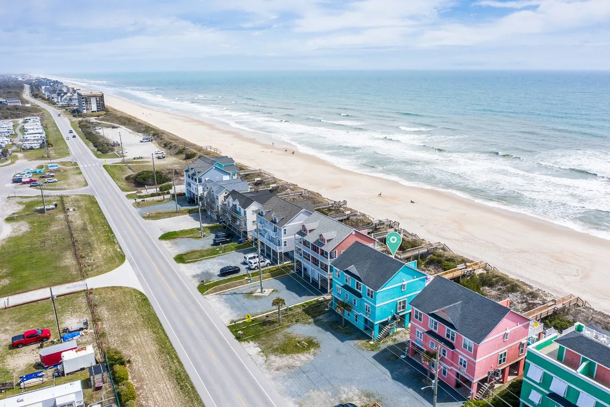 A vibrant exterior view showcases colorful beach houses lined along a wide, sandy beach with gentle ocean waves in the background. The scene is lively, evoking a sense of togetherness and relaxation, perfect for groups enjoying the shore. The open beachfront offers ample space for families to gather, play, and create memories.