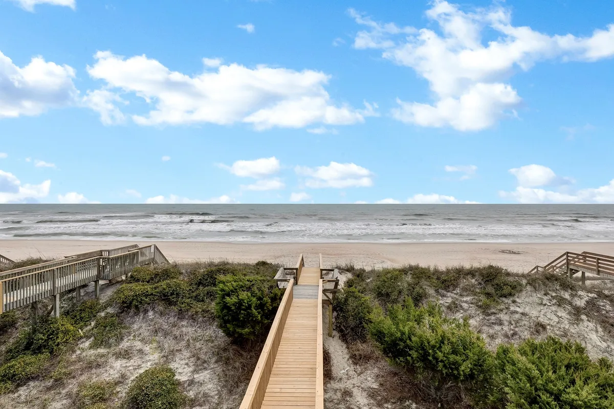 A wooden walkway leads through dune vegetation, opening up to an expansive view of the ocean. The serene setting invites guests and their pets to stroll to the beach and enjoy nature together, highlighting easy access to outdoor adventure.