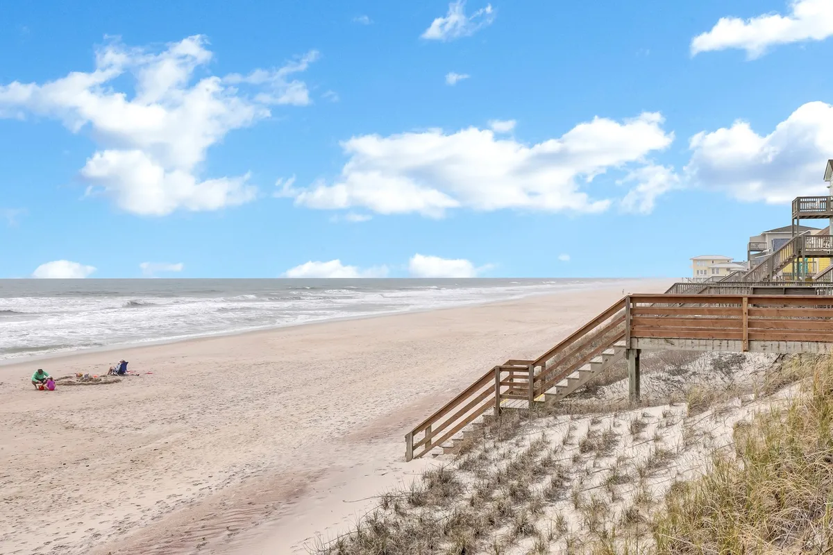 A tranquil stretch of sandy beach meets the gentle surf, with wooden stairs leading down through dune grass. People are seen relaxing on the shore, enjoying the sun, and engaging in classic beach activities like building sandcastles and picnicking.