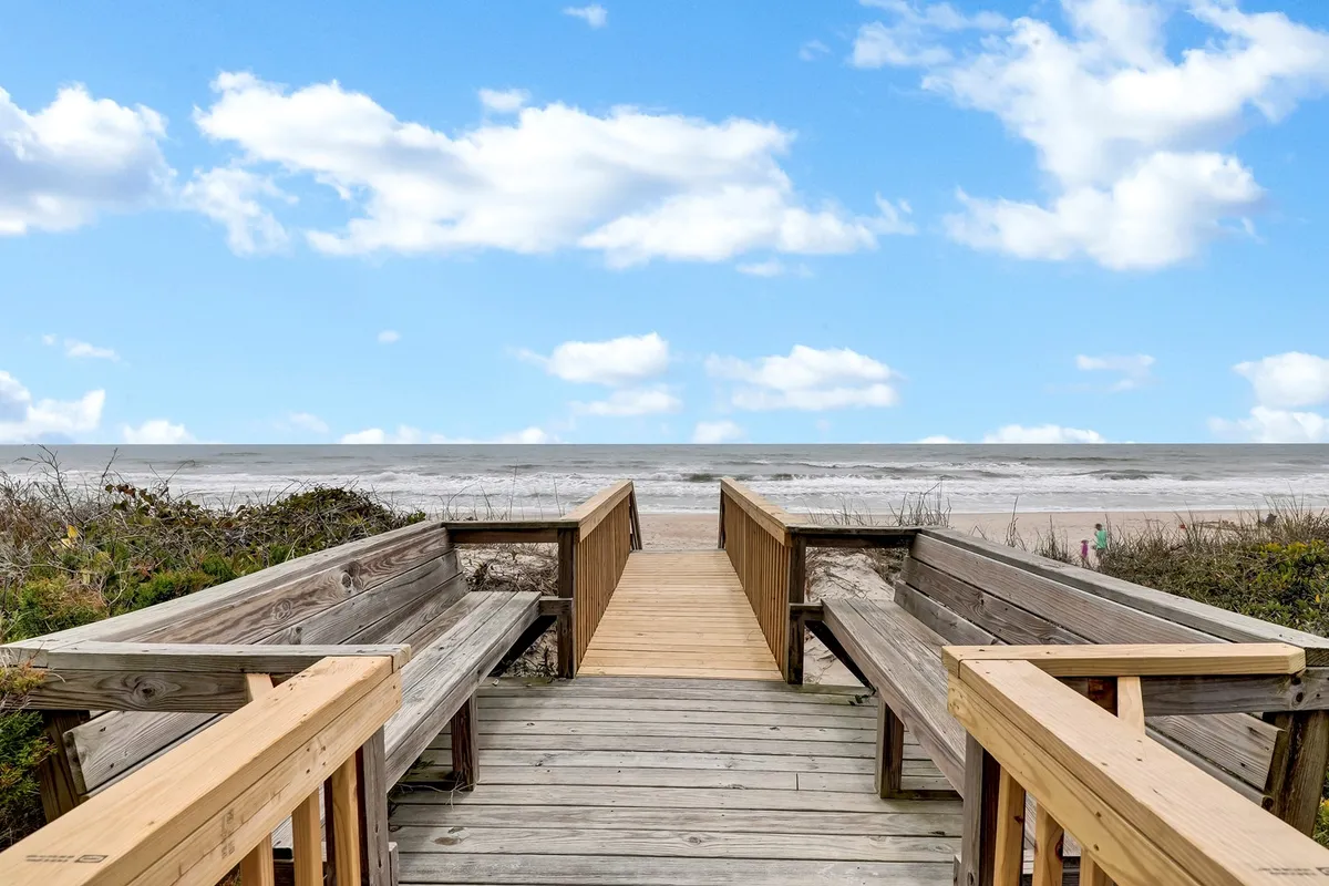 A peaceful wooden walkway leading out to a pristine sandy beach, with blue skies, ocean waves, and benches for taking in the view. The scene feels intimate and offers a sense of anticipation for romantic early mornings or quiet evenings by the sea.