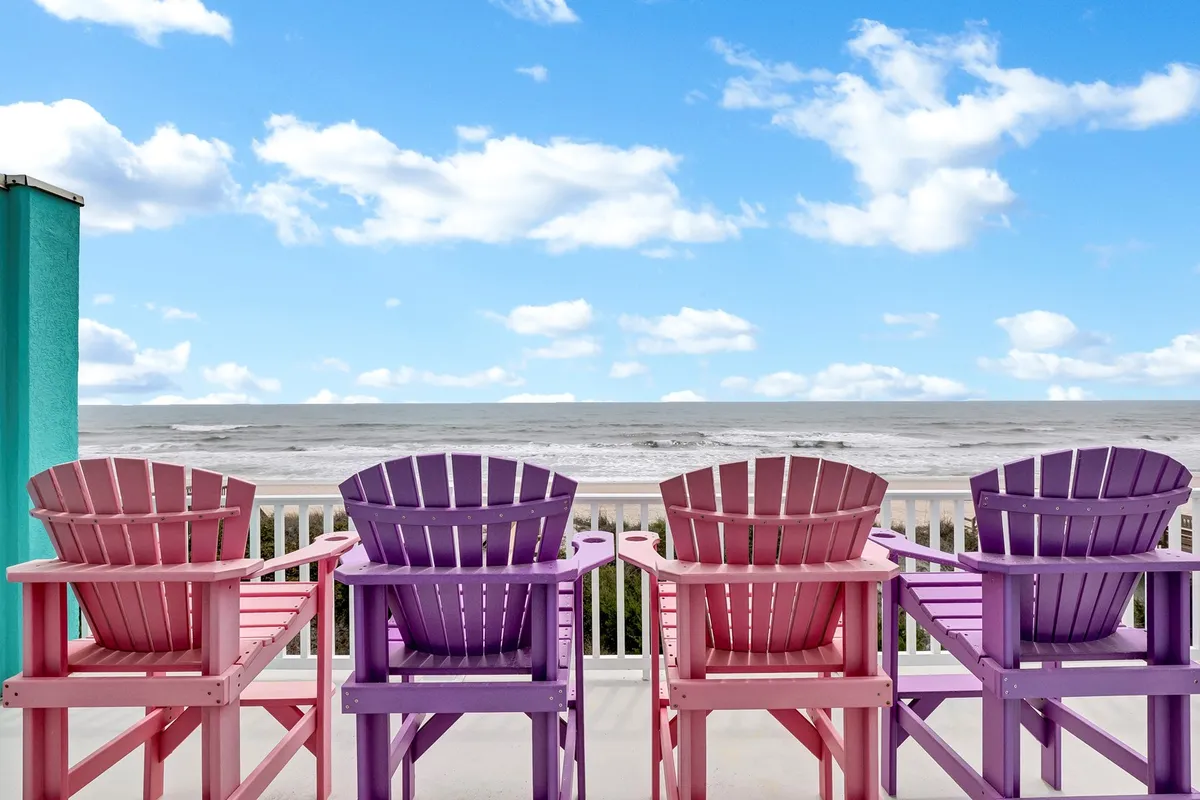 The image shows a row of colorful Adirondack chairs lined up on a spacious deck, overlooking a sweeping ocean view under a dramatic, cloudy sky. The setting evokes a sense of relaxation, inviting guests to unwind and soak in the sounds and sights of the beach, whether enjoying morning coffee or watching the sunset.