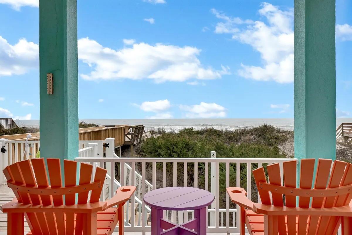 A peaceful porch setting with two colorful Adirondack chairs and a small purple table facing an ocean view. The space is ideal for relaxing, enjoying the fresh air, and provides easy access from indoors to out—perfect for quick pet walks or unwinding after a sandy stroll.
