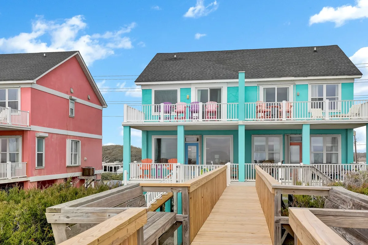 Colorful balconies and a cheerful wooden walkway frame brightly painted houses near the beach. The scene hints at community, outdoor relaxation, and vibrant beach life, with plenty of outdoor space for families and their pets to enjoy time together.