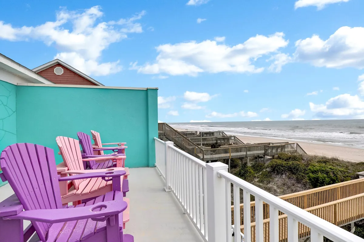A cheerful exterior balcony scene featuring pink Adirondack chairs set against a turquoise wall, with a breathtaking ocean view beyond. The setup feels casual and inviting, encouraging socializing and relaxation while watching sunsets or enjoying morning coffee together.