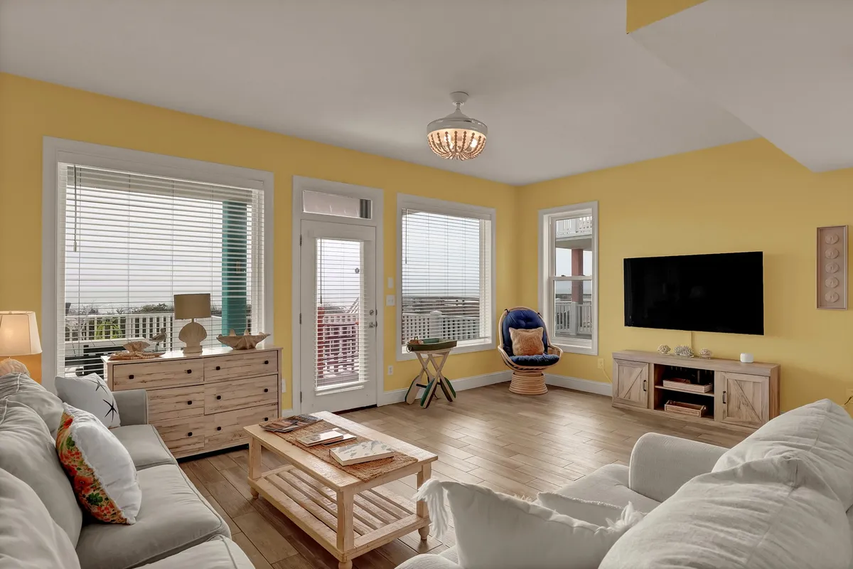 A sunlit living room featuring comfortable seating, yellow walls, a wooden coffee table, large windows, and a TV. The expansive windows reveal ocean views, and the space feels open, airy, and ideal for gatherings or relaxing.