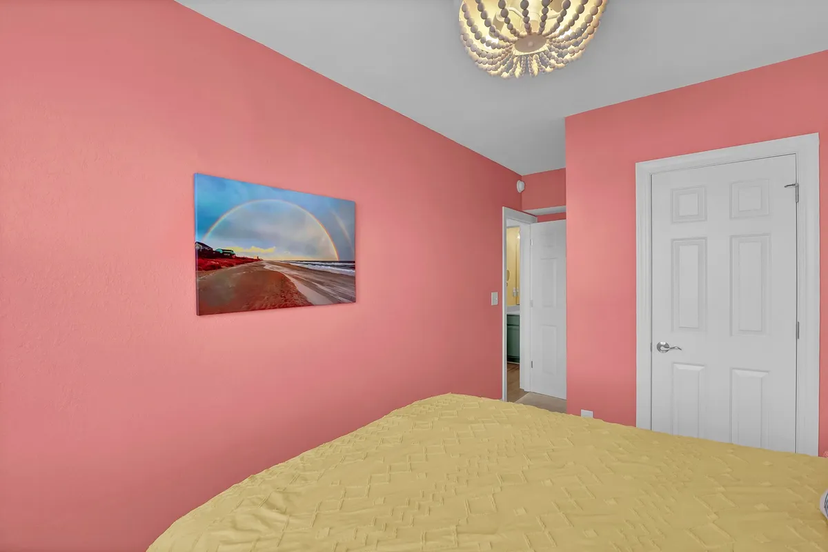 A cheerful bedroom with a pink wall, yellow bedding, ocean landscape artwork, and a sparkling chandelier. The room feels bright and welcoming, ideal for guests or family members seeking a cozy retreat.