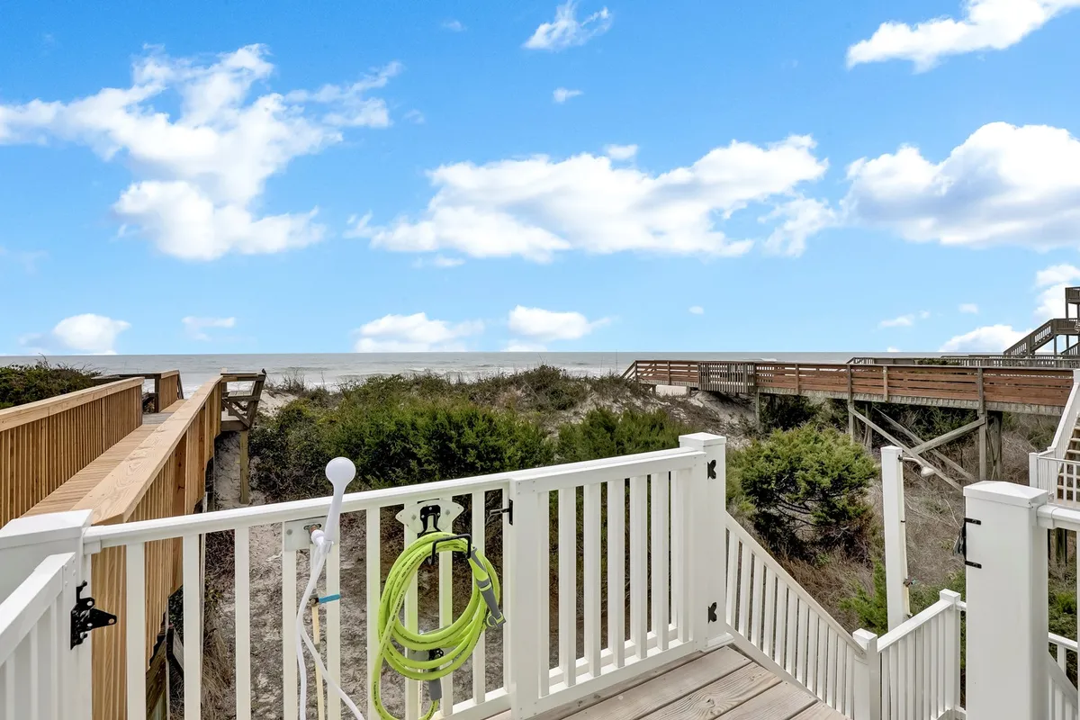 A serene wooden walkway stretches through lush green vegetation toward the ocean, framed by a classic white railing. The view highlights easy access to the beach and invites exploration and family gatherings.