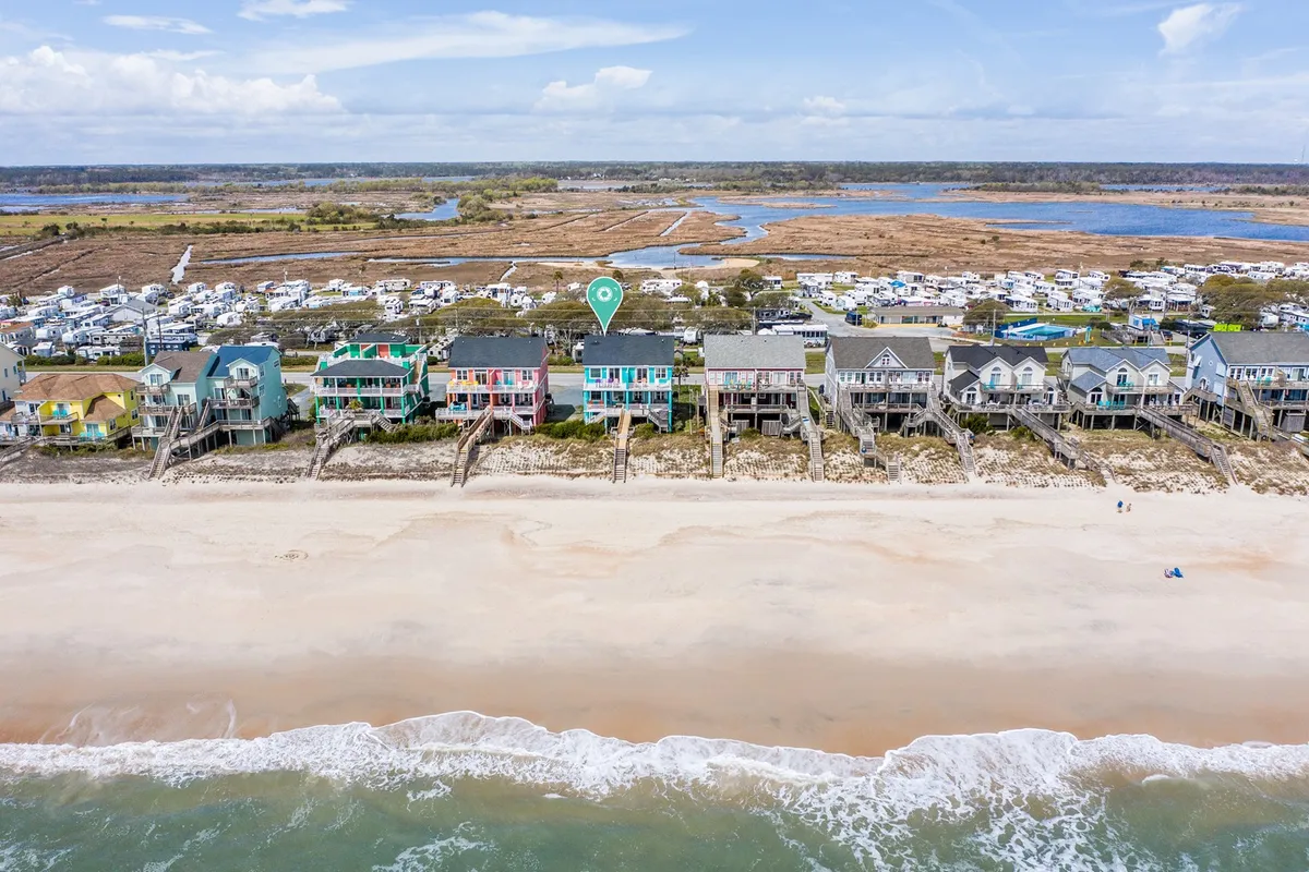 A cheerful exterior scene of colorful beach houses lining a sandy shoreline, with the ocean and marshland beyond, under a bright blue sky. The setting is sunny, open, and welcoming, inviting guests to enjoy nature and sustainable travel.