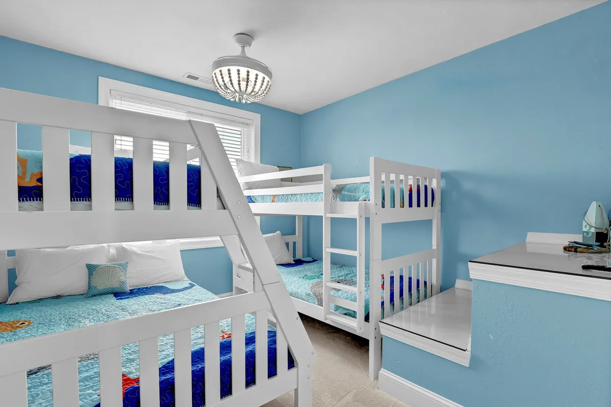 A playful and inviting kids’ bunk room with blue walls, built-in bunk beds, and a whimsical chandelier overhead. The space is bright and cheerful, perfect for children to unwind after a day outside.