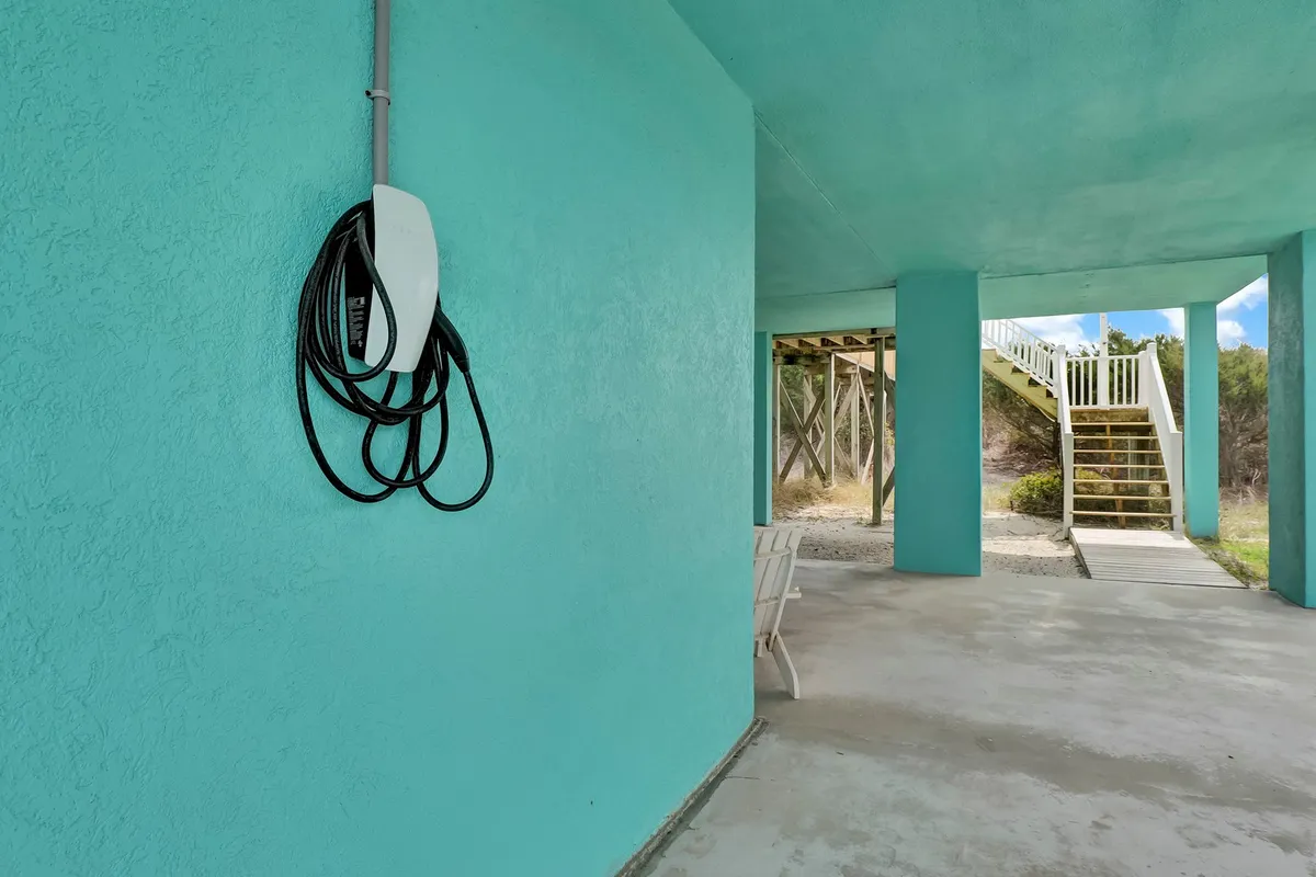 This exterior image highlights a turquoise wall with an electric vehicle charger prominently visible beside a staircase that leads outdoors. There is a white chair positioned to suggest a welcoming spot for relaxing just outside the entrance, framed by natural light and open air.